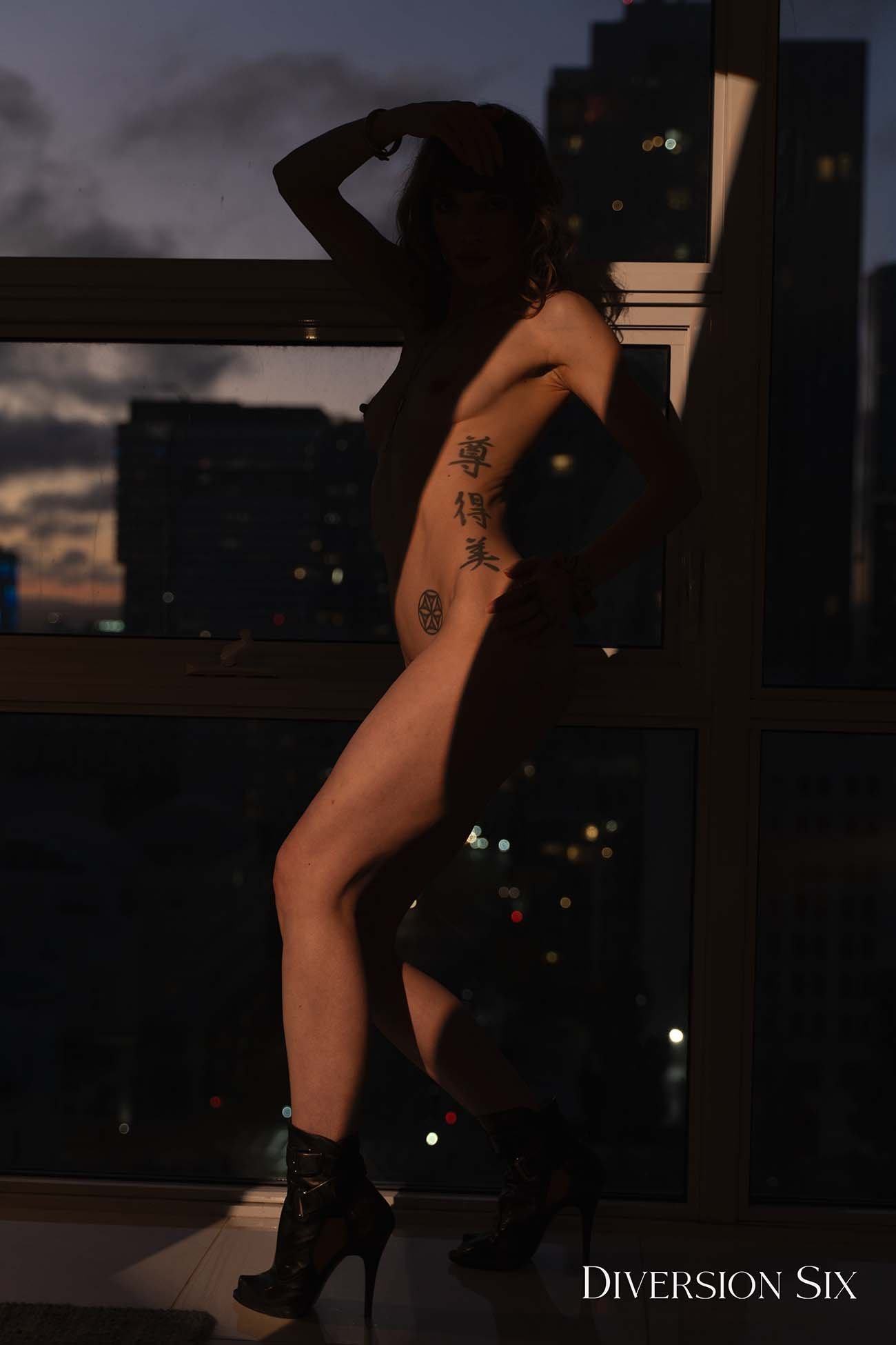 A nude blond model in the dark in a sexy pose with the a ray of light across her body and the city lights in the background